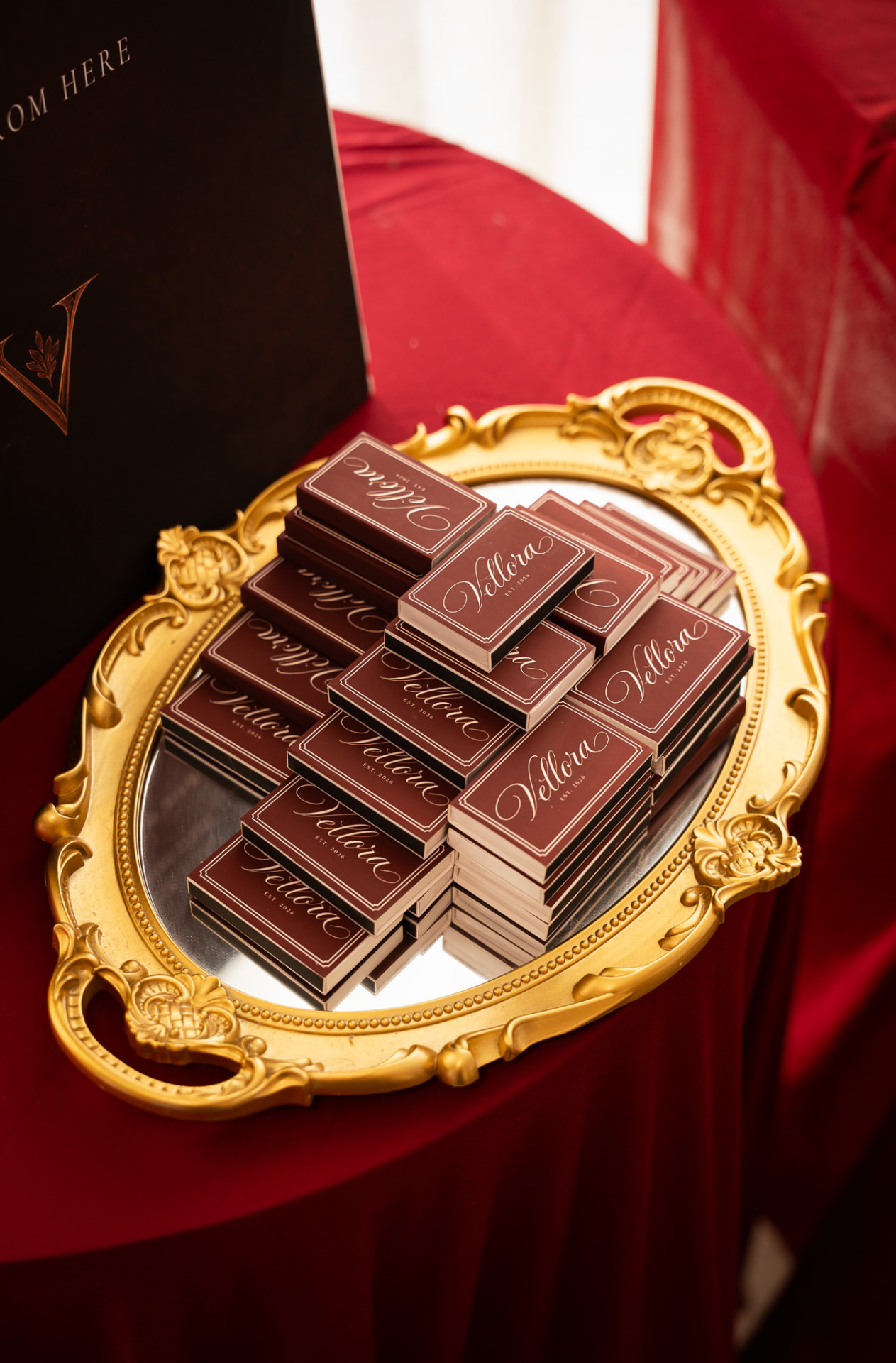 An ornate gold tray on a red clothed table holding stacks of wine coloured Vellora matchboxes beside the house sign