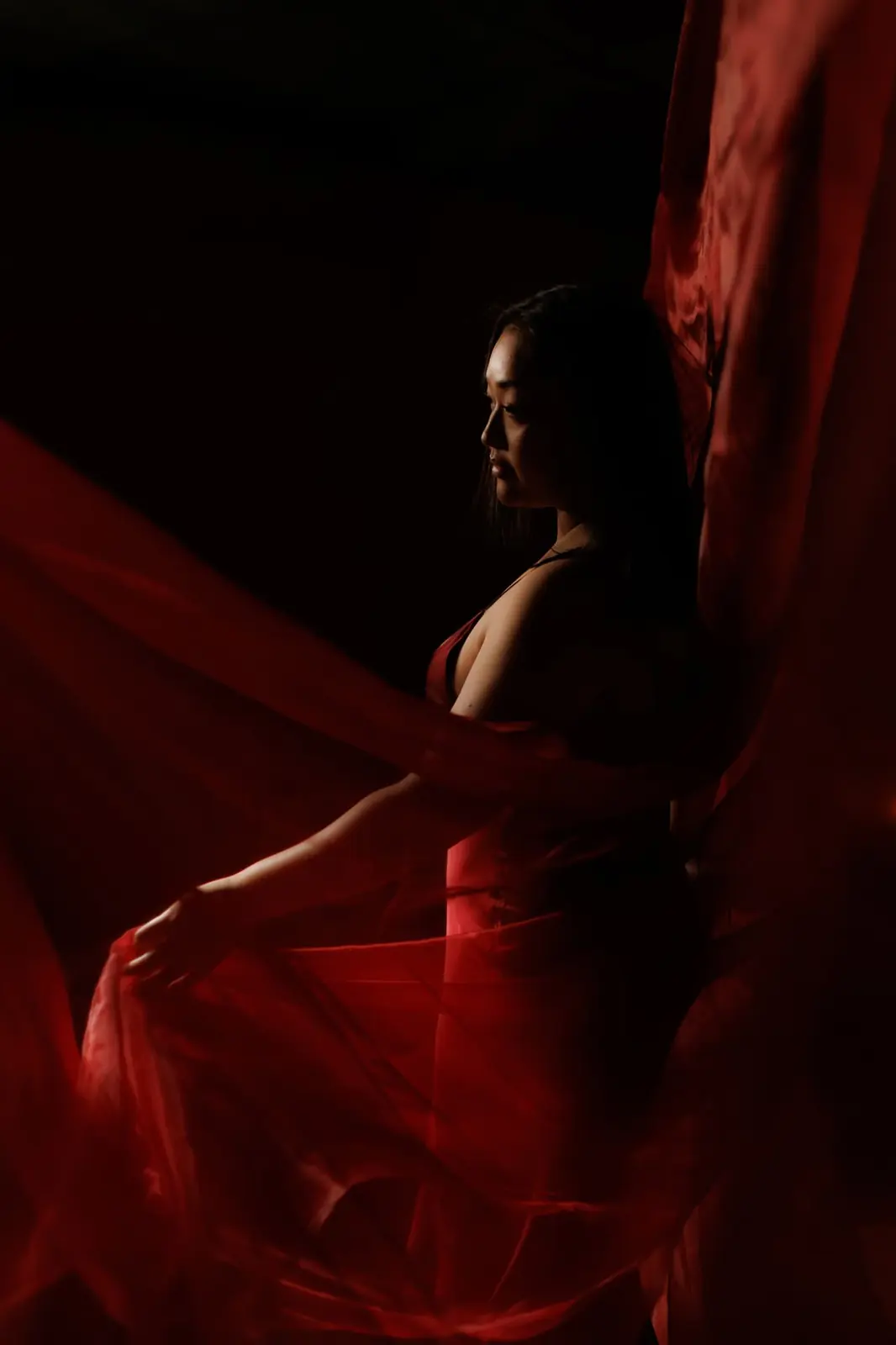 A woman in profile drawing a length of red chiffon around her against a black backdrop