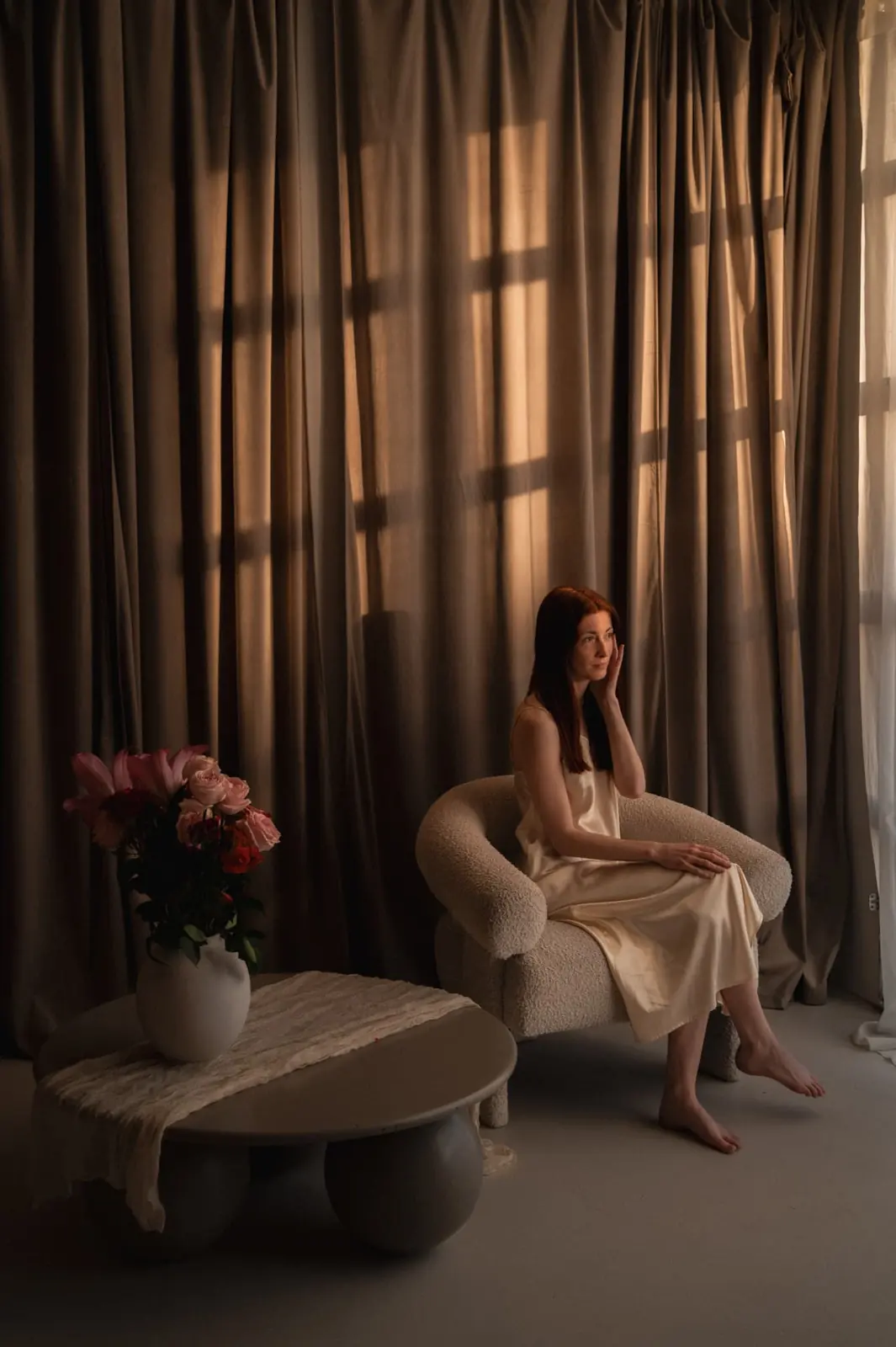 A woman in champagne silk seated in a boucle chair as late window light falls across floor length curtains and roses in a pale vase