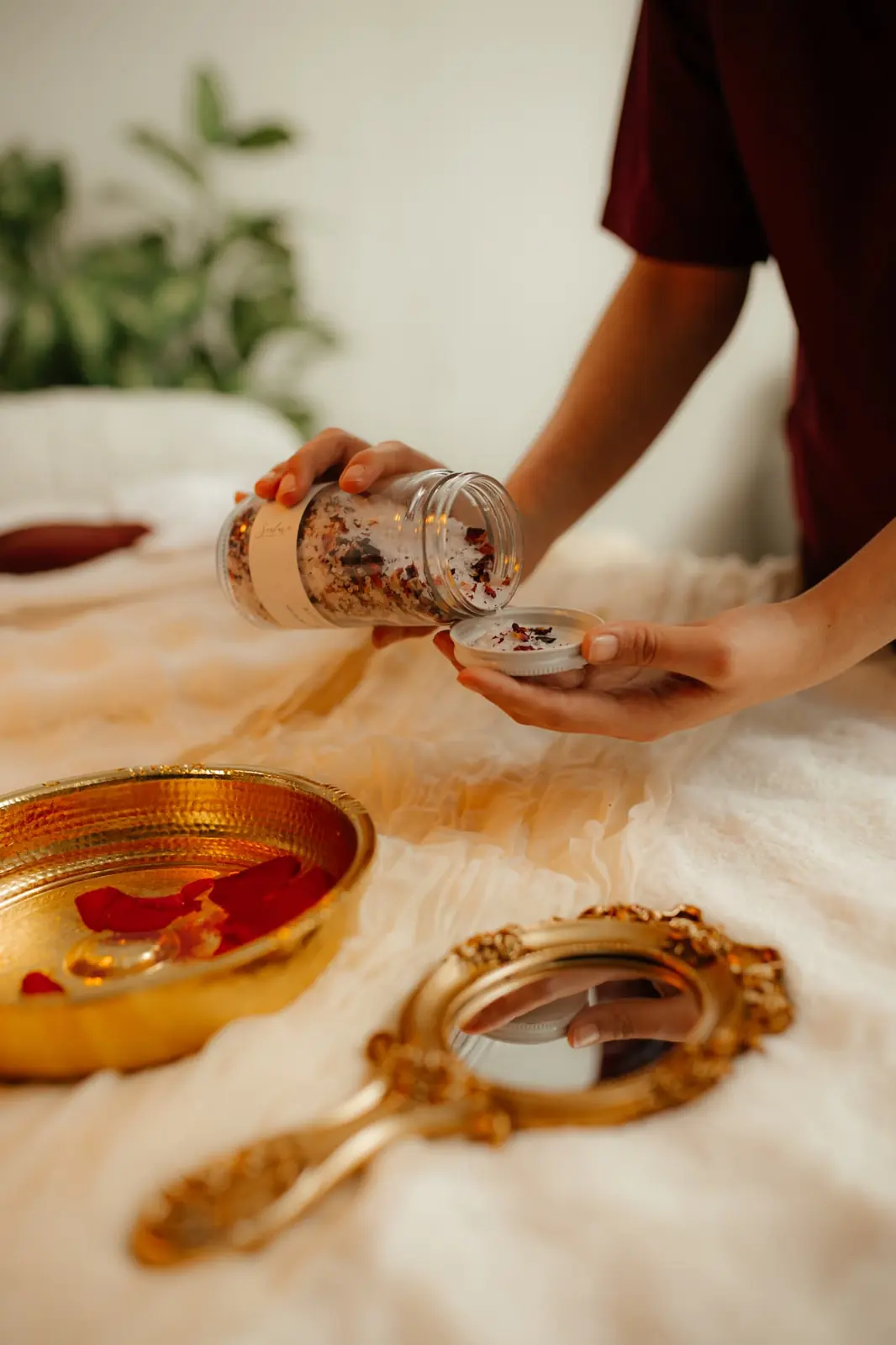 A practitioner tipping rose petal bath salts from a glass jar into its lid beside a hammered gold bowl of water and a gold hand mirror