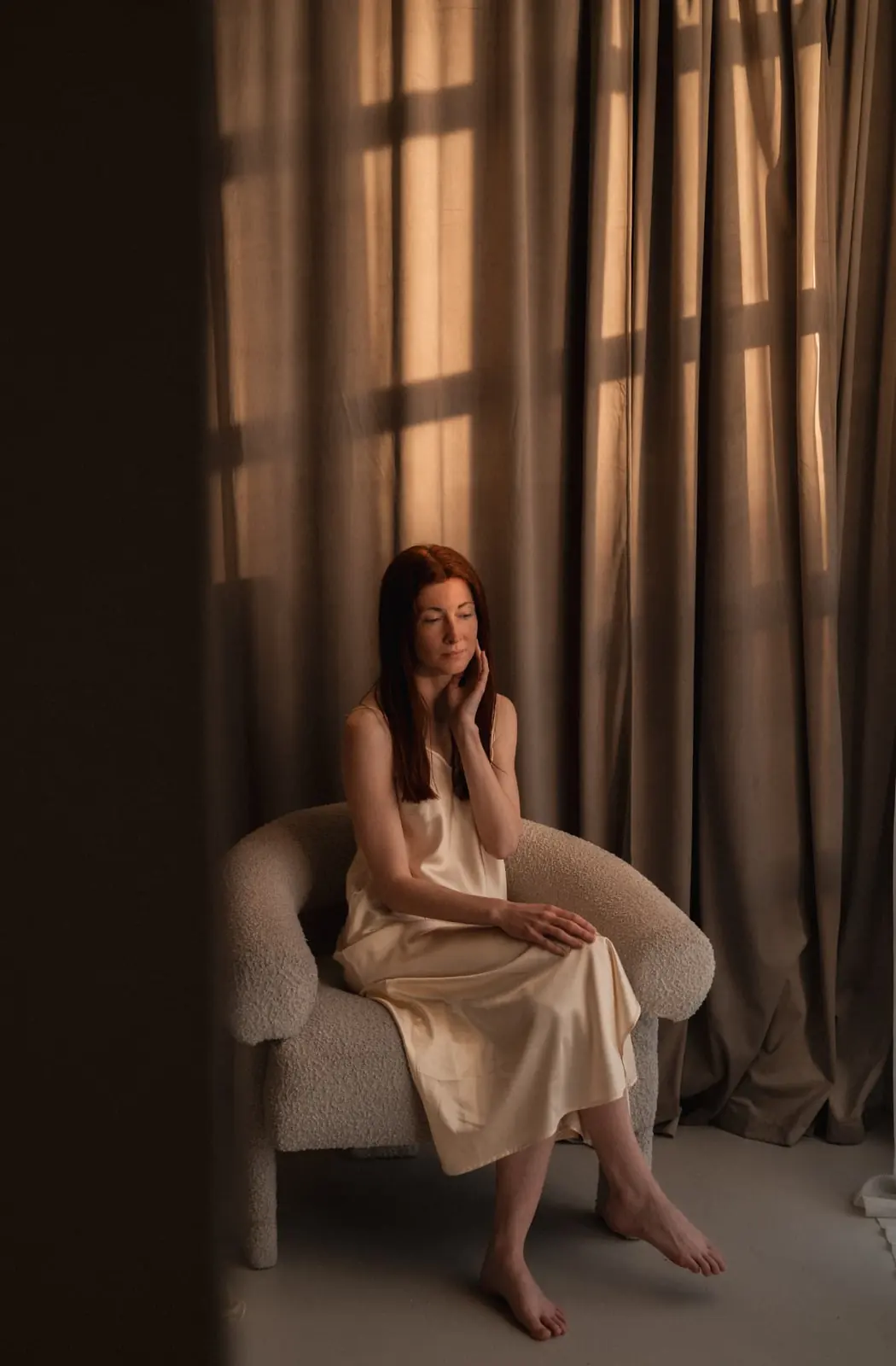 A woman in champagne silk resting a hand against her jaw in a boucle chair, window shadows across the curtain behind her