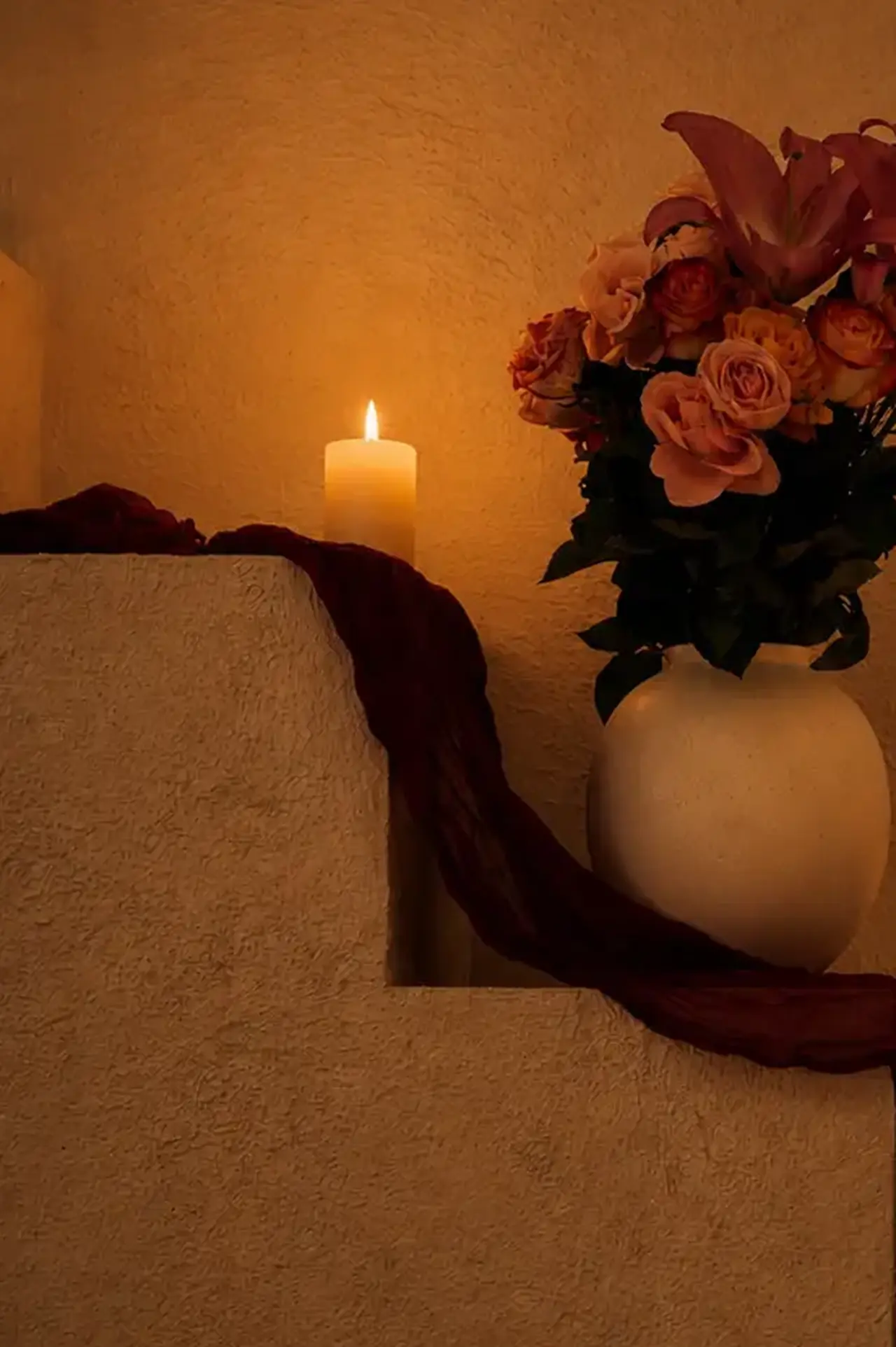 Wine-coloured gauze folded over plaster steps beside a burning pillar candle and roses in a pale vase