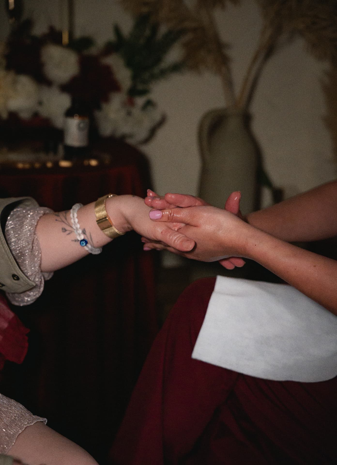 A practitioner hands working warm botanical oil into a guest hand in a candlelit room of roses and dark red drapes