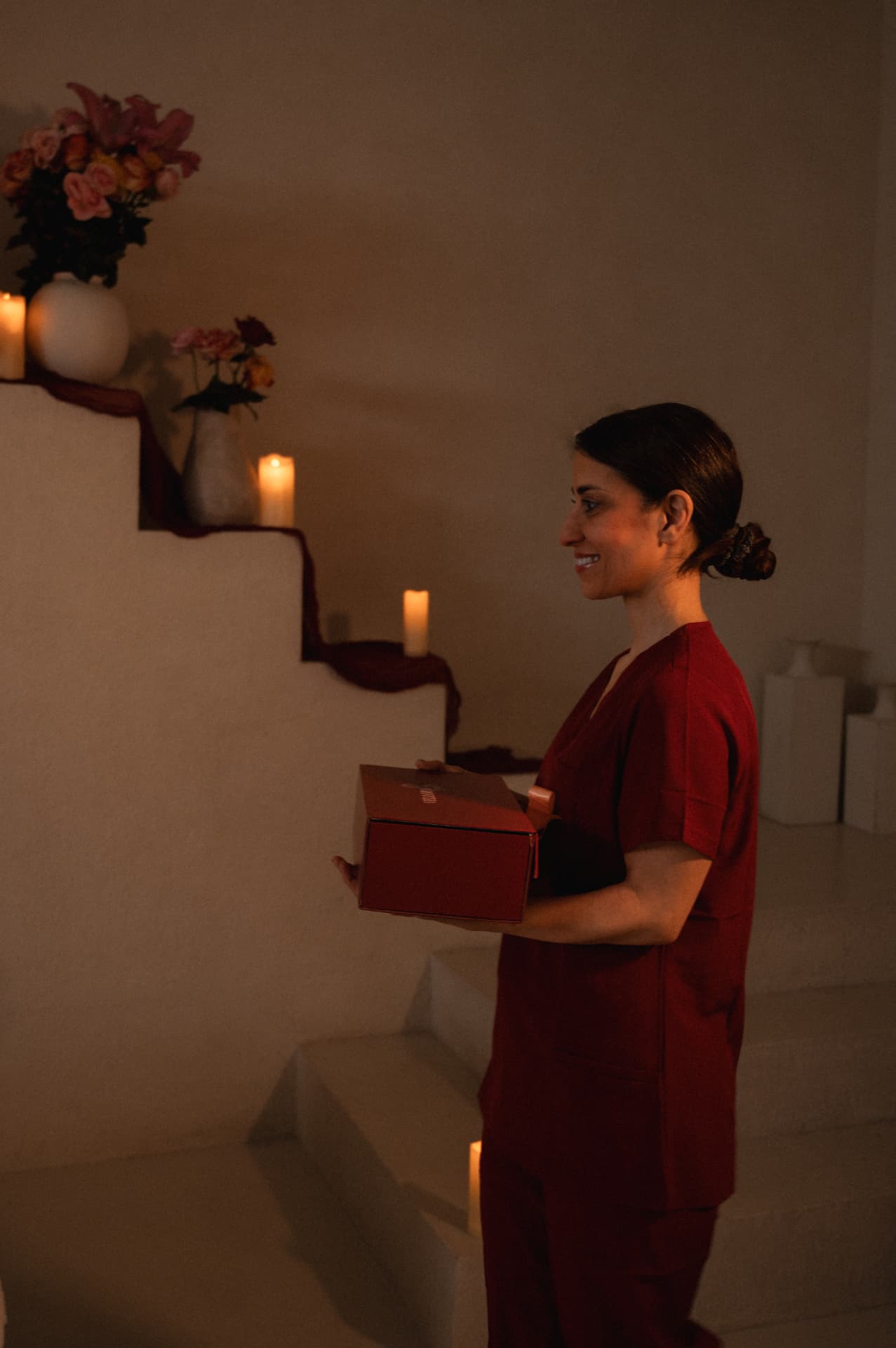 A practitioner in wine linen passing a deep red keepsake box to a seated woman beside candlelit plaster steps