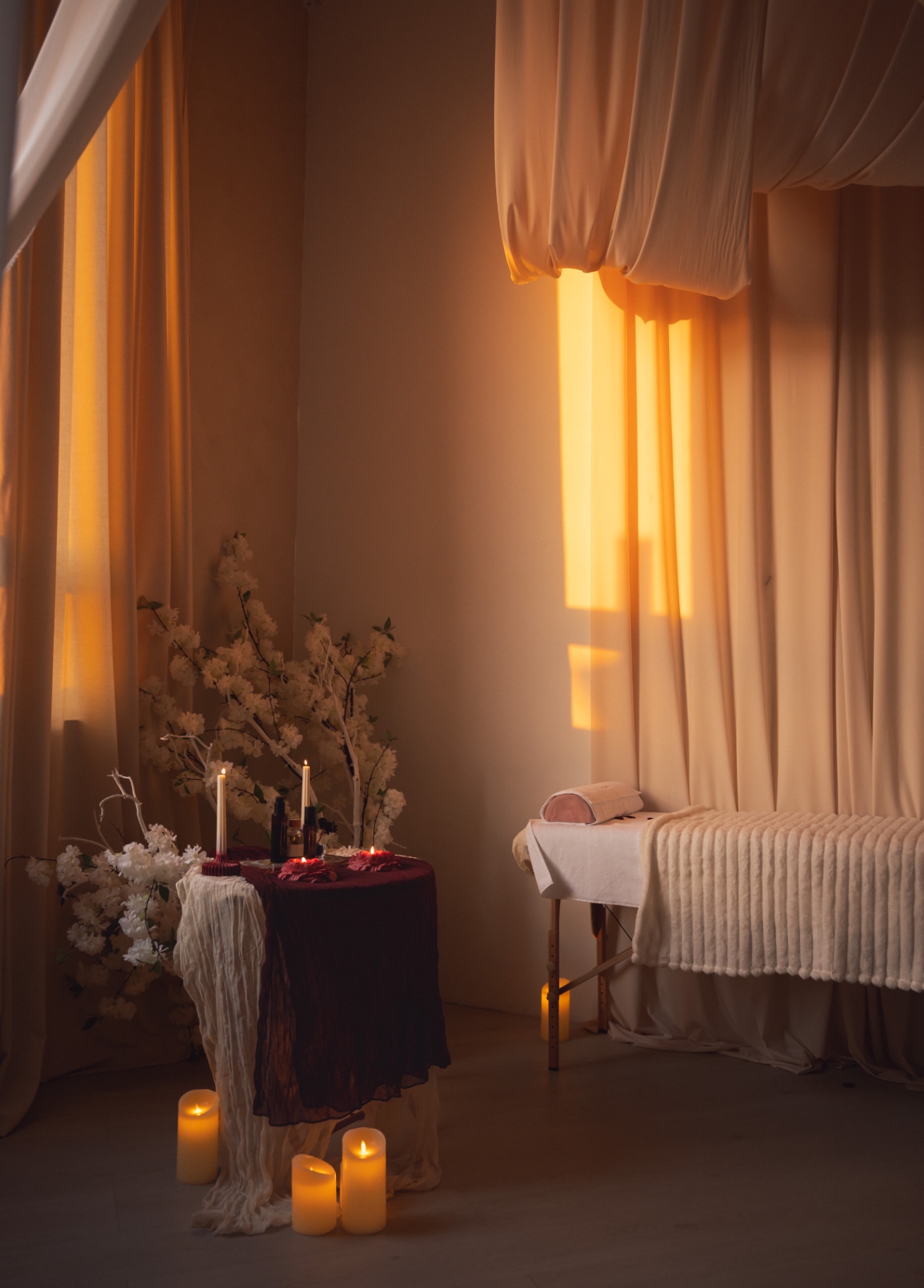 A small draped table holding lit pillar candles and glass bottles beside a low massage bed, warm light falling through cream curtains