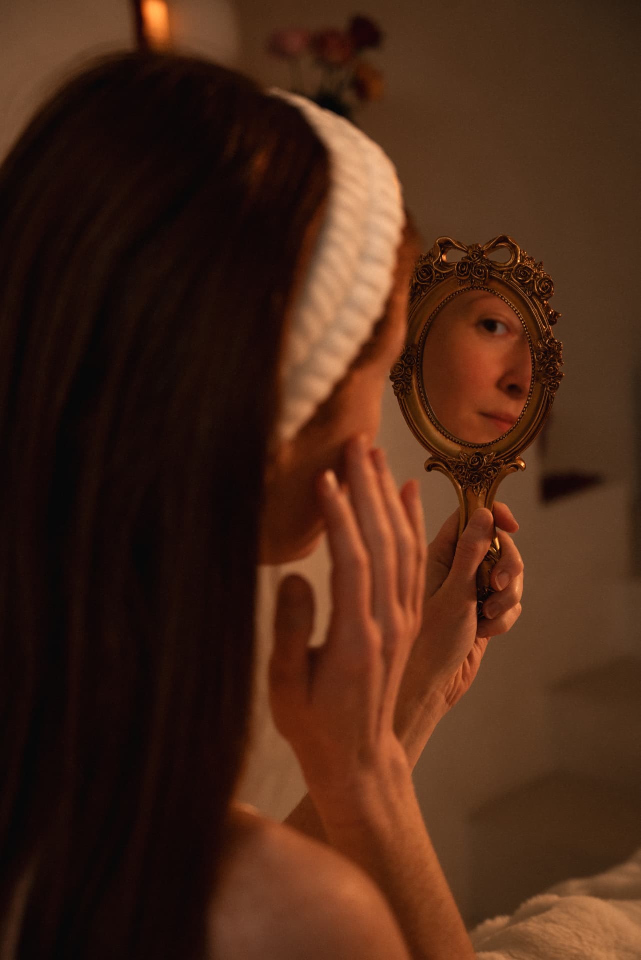 A woman reflected in a warm gilt mirror, her shoulders loose in the low light of a prepared room
