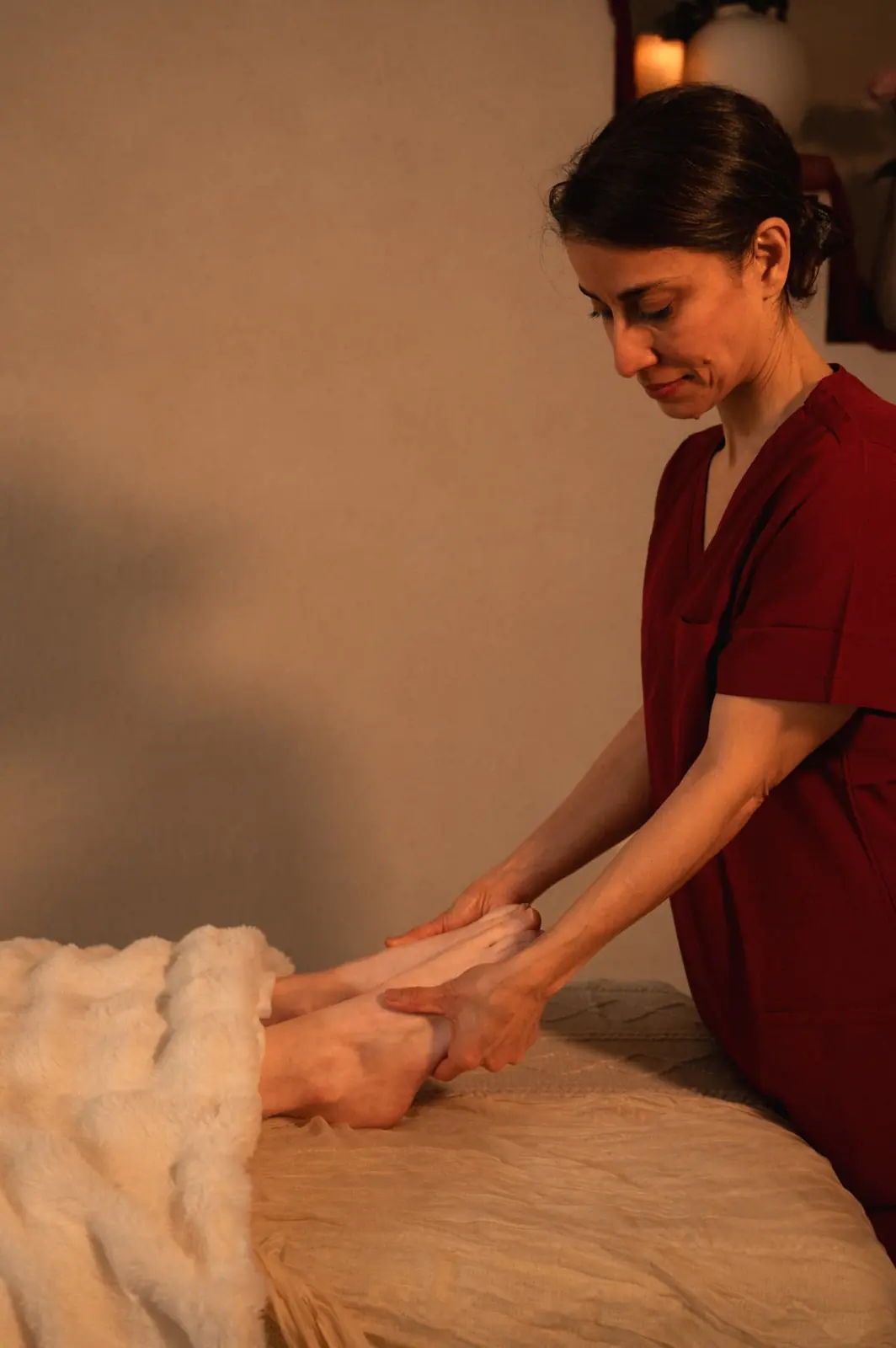 A practitioner in wine scrubs cradling a woman feet at the end of the table under a cream blanket