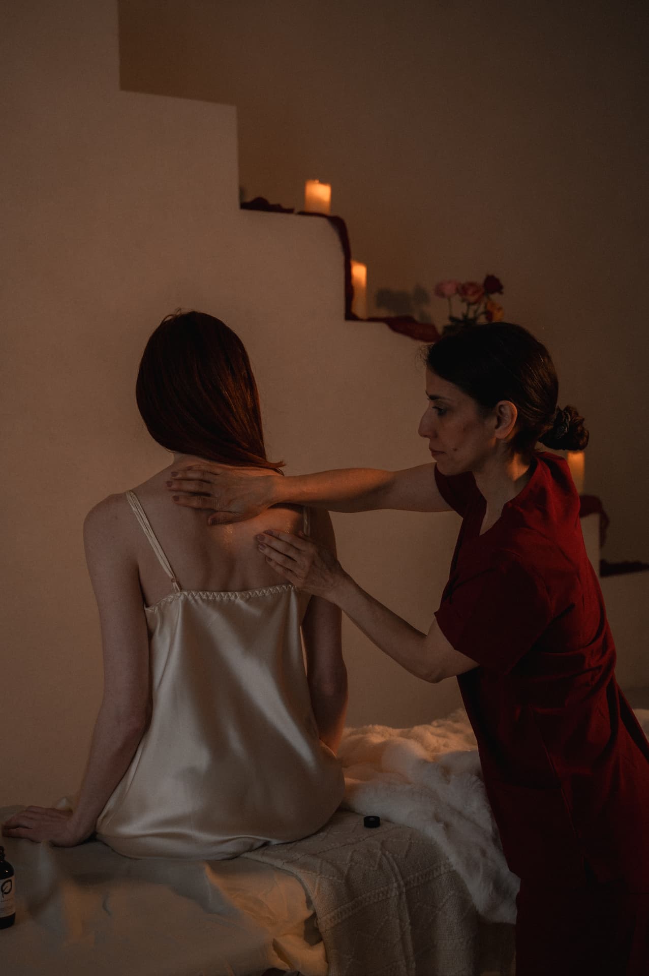 A woman seated upright in cream silk while a practitioner in red rests both hands across her shoulders in candlelight