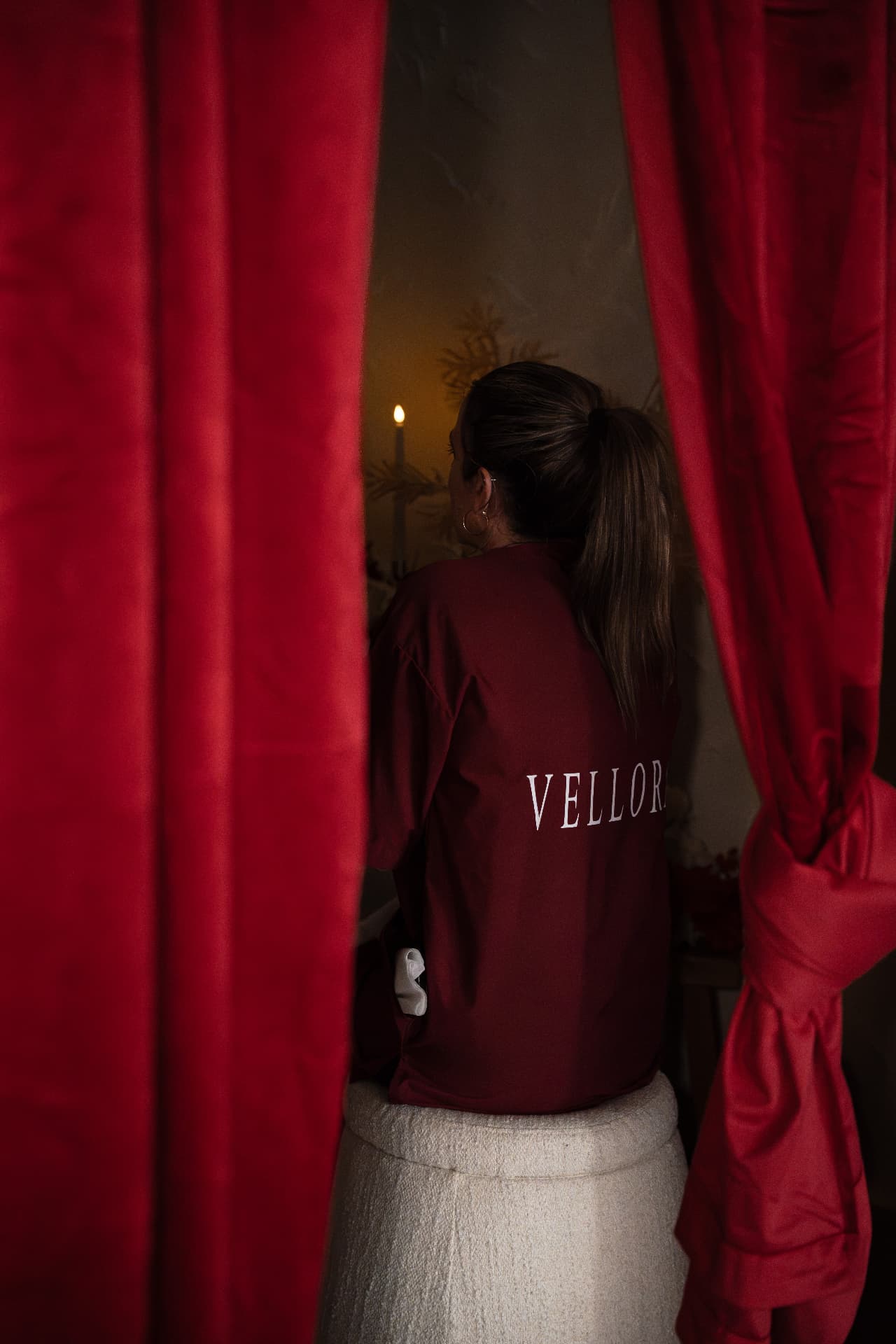 A practitioner of the house in a wine Vellora shirt seated between drawn red velvet curtains, a single candle lit behind her