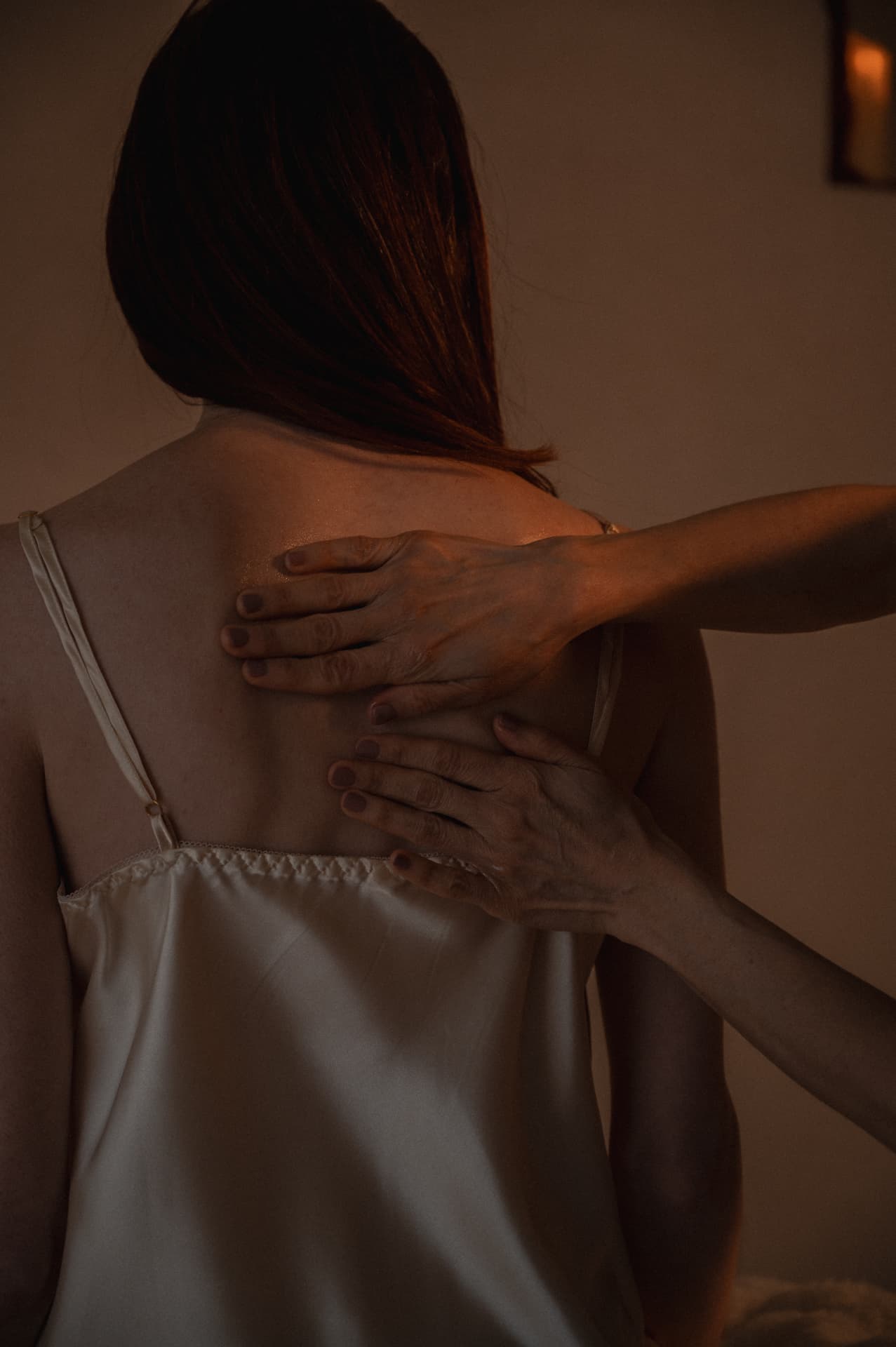 Two hands pressing with slow weight along a woman upper back in a warm, dim candlelit room