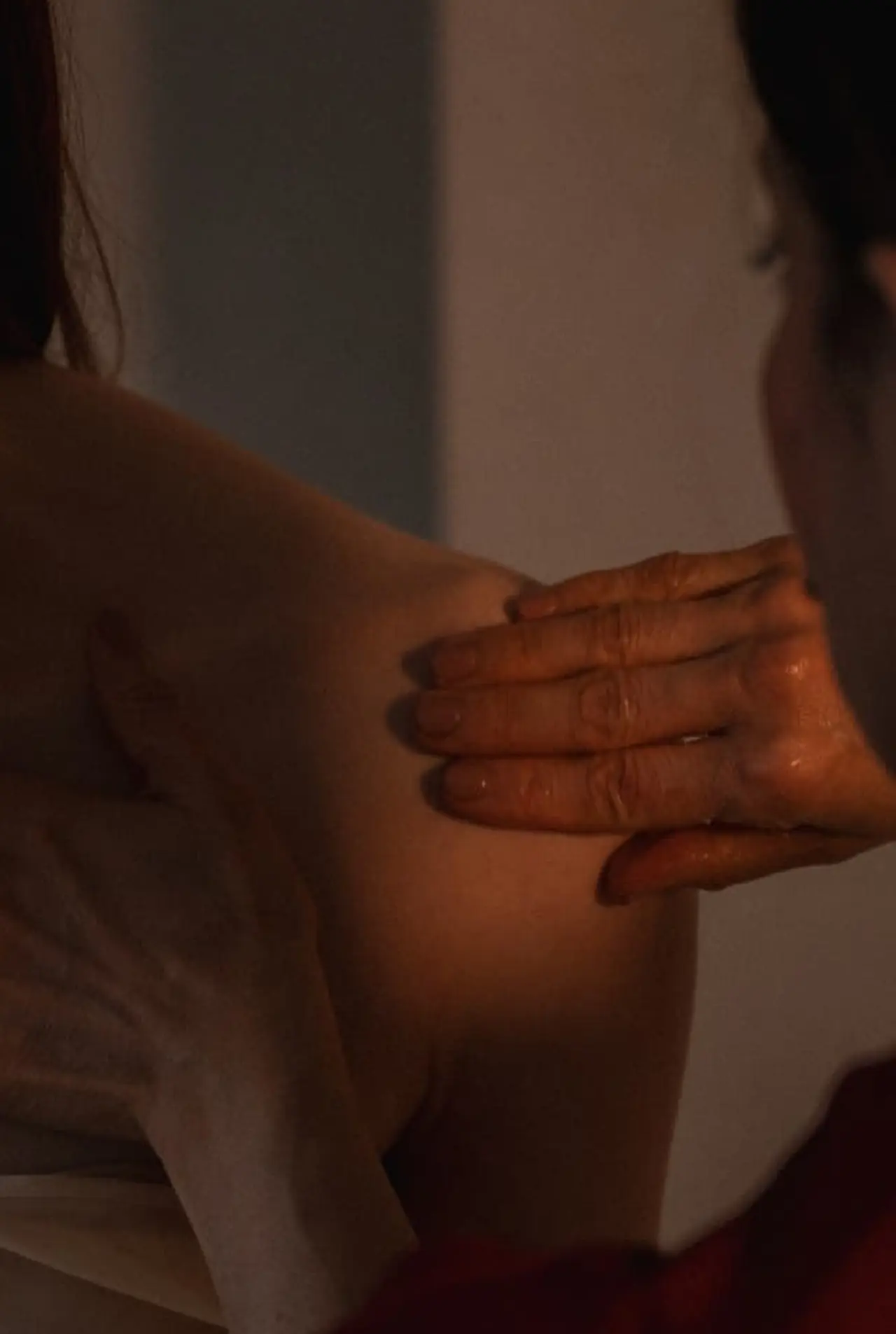 A practitioner hand working warm oil slowly across a woman bare shoulder in low light