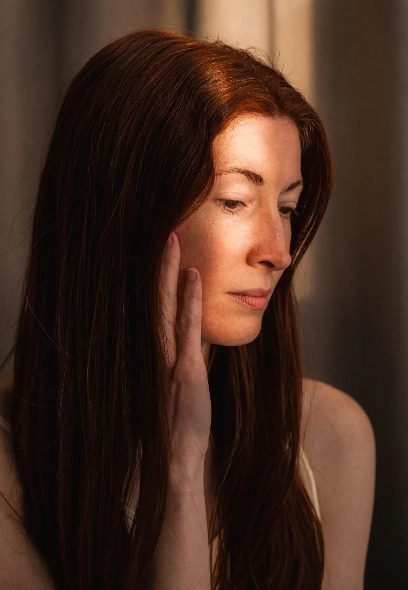 A woman with long auburn hair resting her cheek against her hand in warm low light